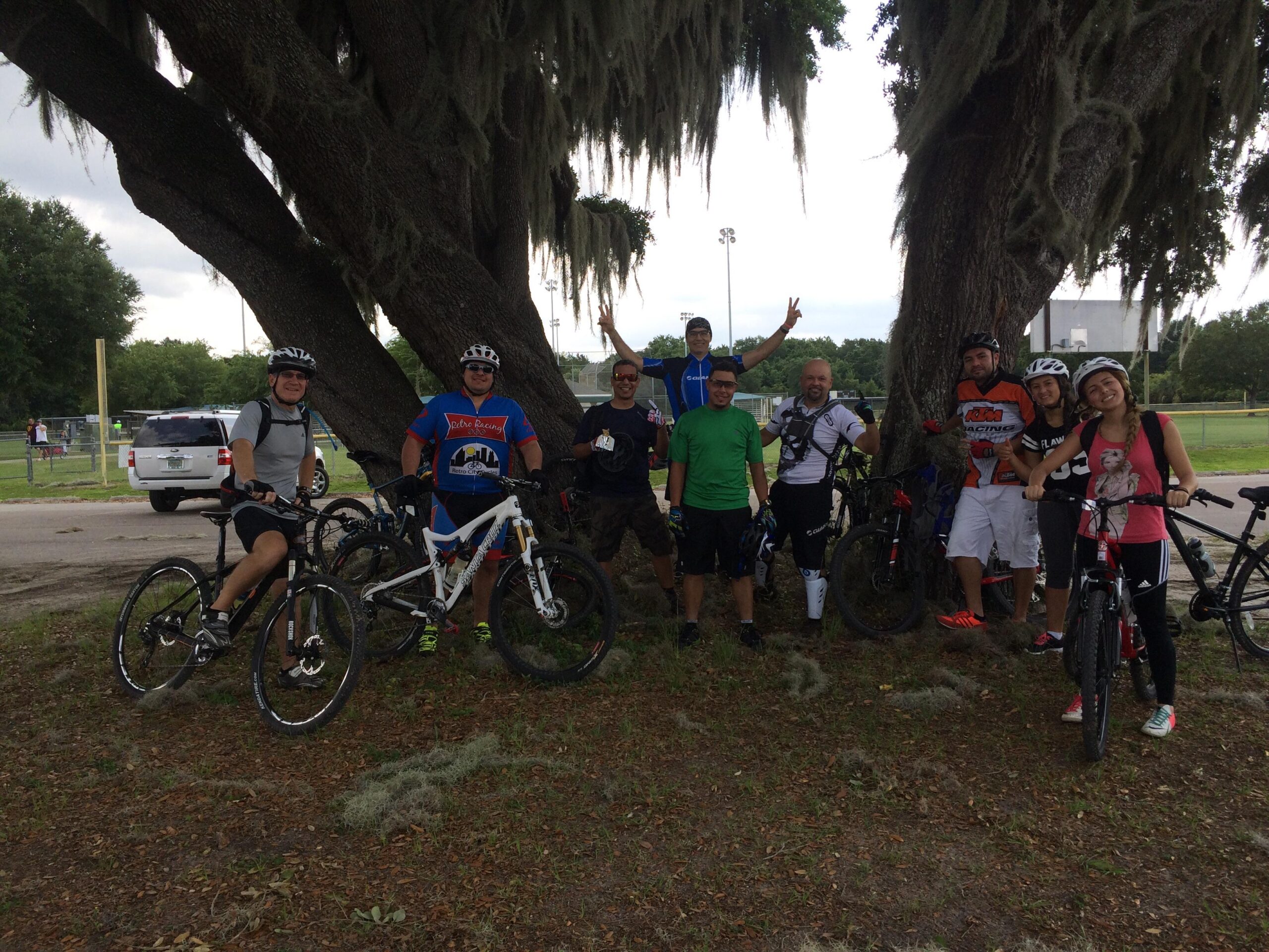 A group of eight people enjoying a break during a mountain biking outing, standing beside their bicycles under a large tree adorned with Spanish moss. The individuals, wearing helmets and biking gear, are smiling and posing for the photo, with some holding up peace signs. In the background, a park setting with a sports field and parked vehicles can be seen. Rollercoaster mountain bike trail.