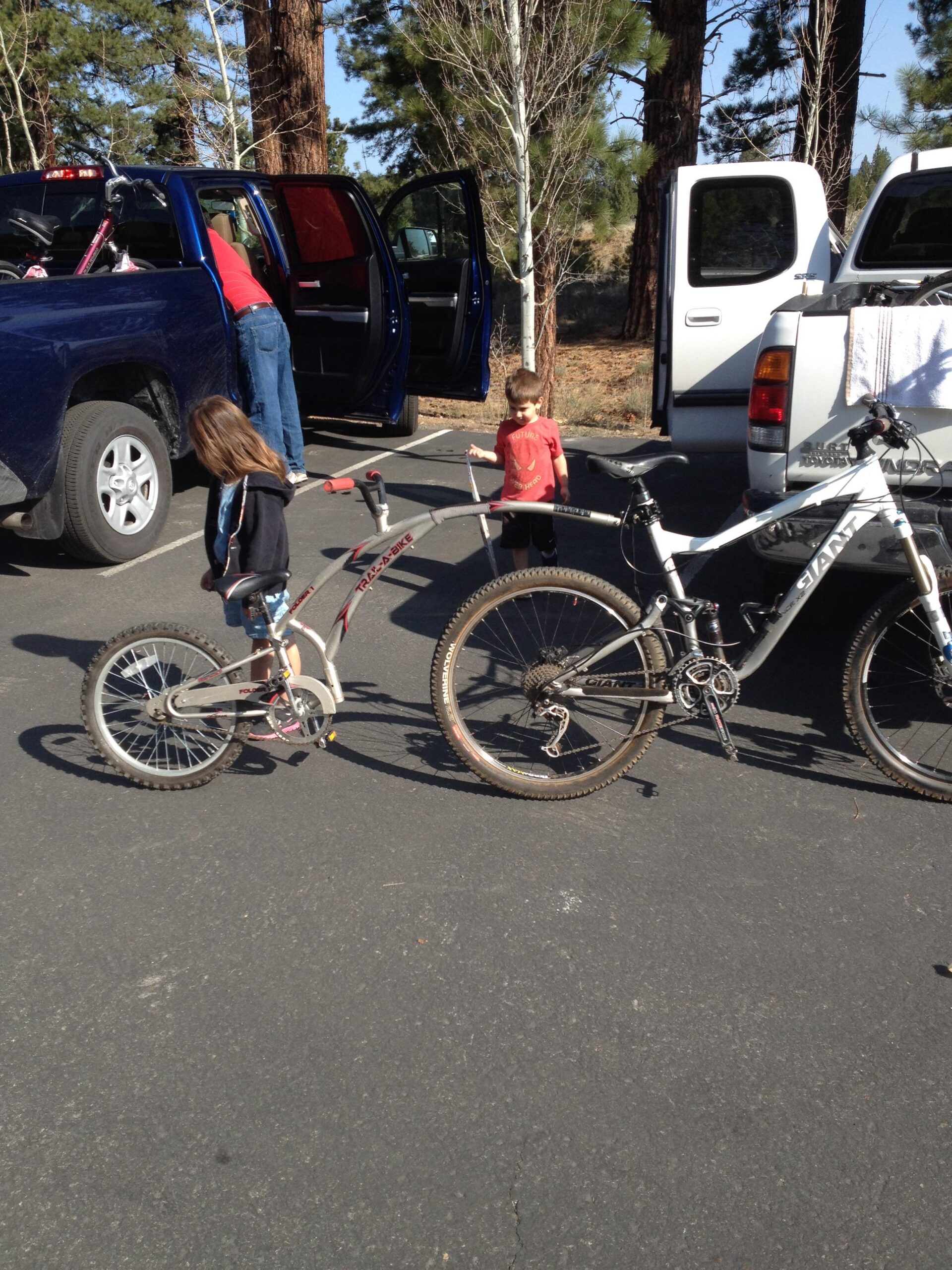Giant Trance: A child walking beside a bike while another child plays nearby. A blue truck with its doors open is in the background, and a second white truck is parked nearby. The scene takes place in a wooded area with trees in the background.