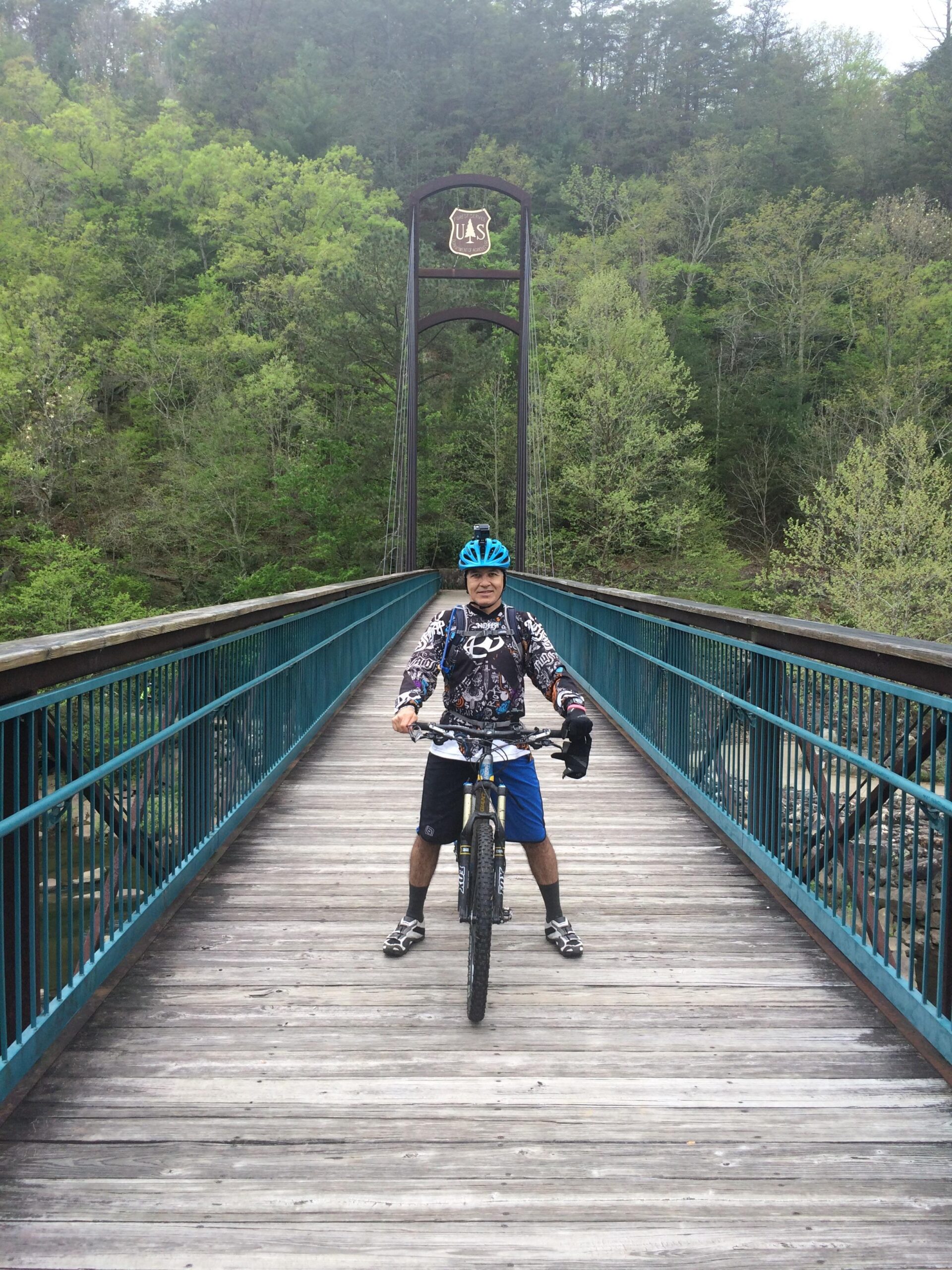 A person wearing a blue helmet and biking gear stands on a wooden bridge, holding a mountain bike. Lush green trees surround the bridge, and a U.S. Forest Service sign is visible above. The scene captures a moment in a natural outdoor setting ideal for biking. Tanasi Trail System mountain bike trail.