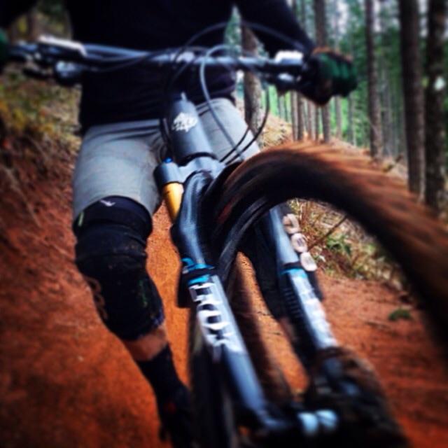 A close-up view of a mountain bike handlebar and front wheel, with a rider's legs visible in biking gear, navigating a dirt trail surrounded by tall trees. The image captures the motion and excitement of biking in a natural setting. Post Canyon mountain bike trail.