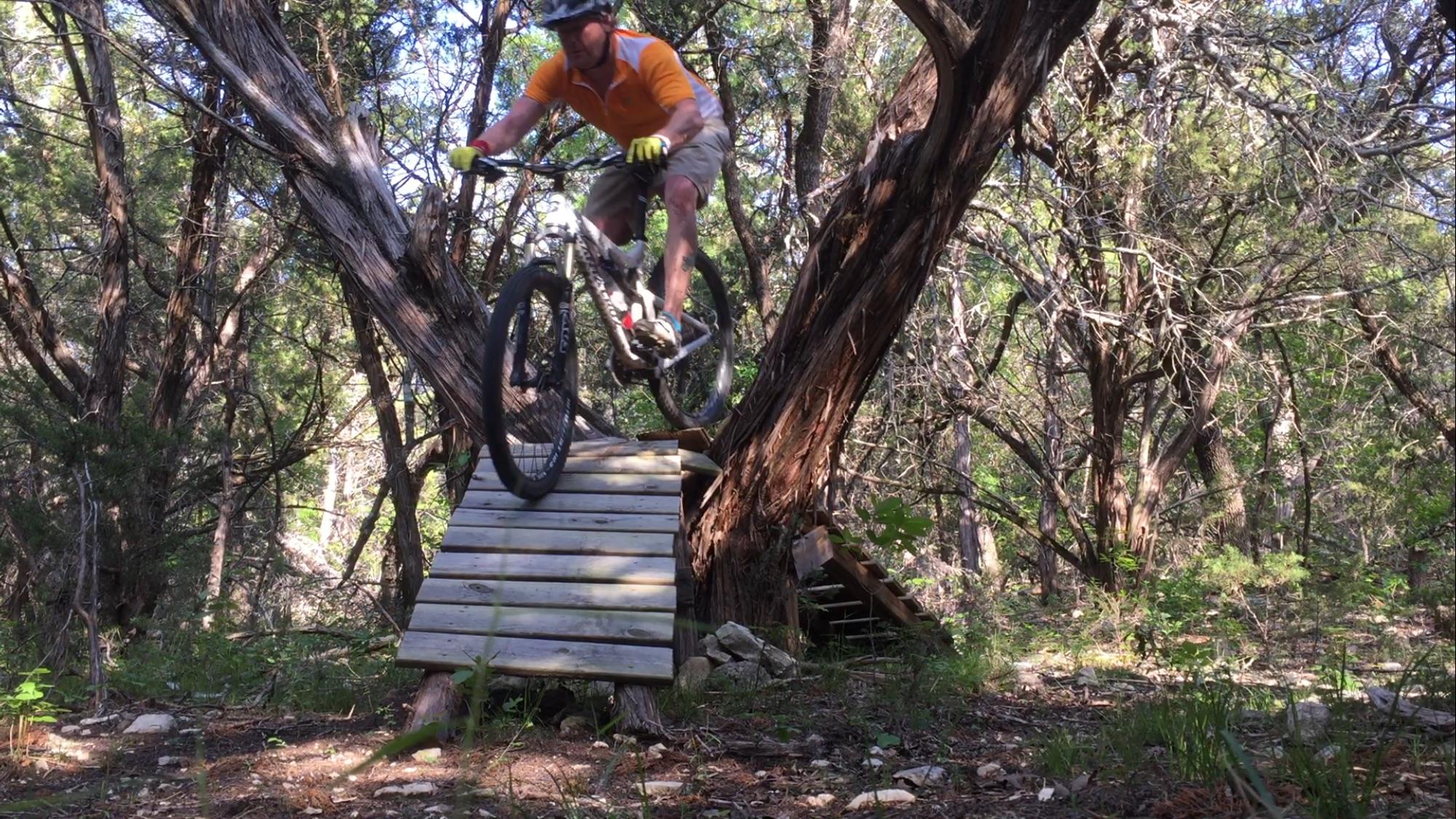 A mountain biker in an orange shirt and shorts is mid-jump over a wooden ramp in a wooded area, surrounded by trees and greenery. Blora mountain bike trail.