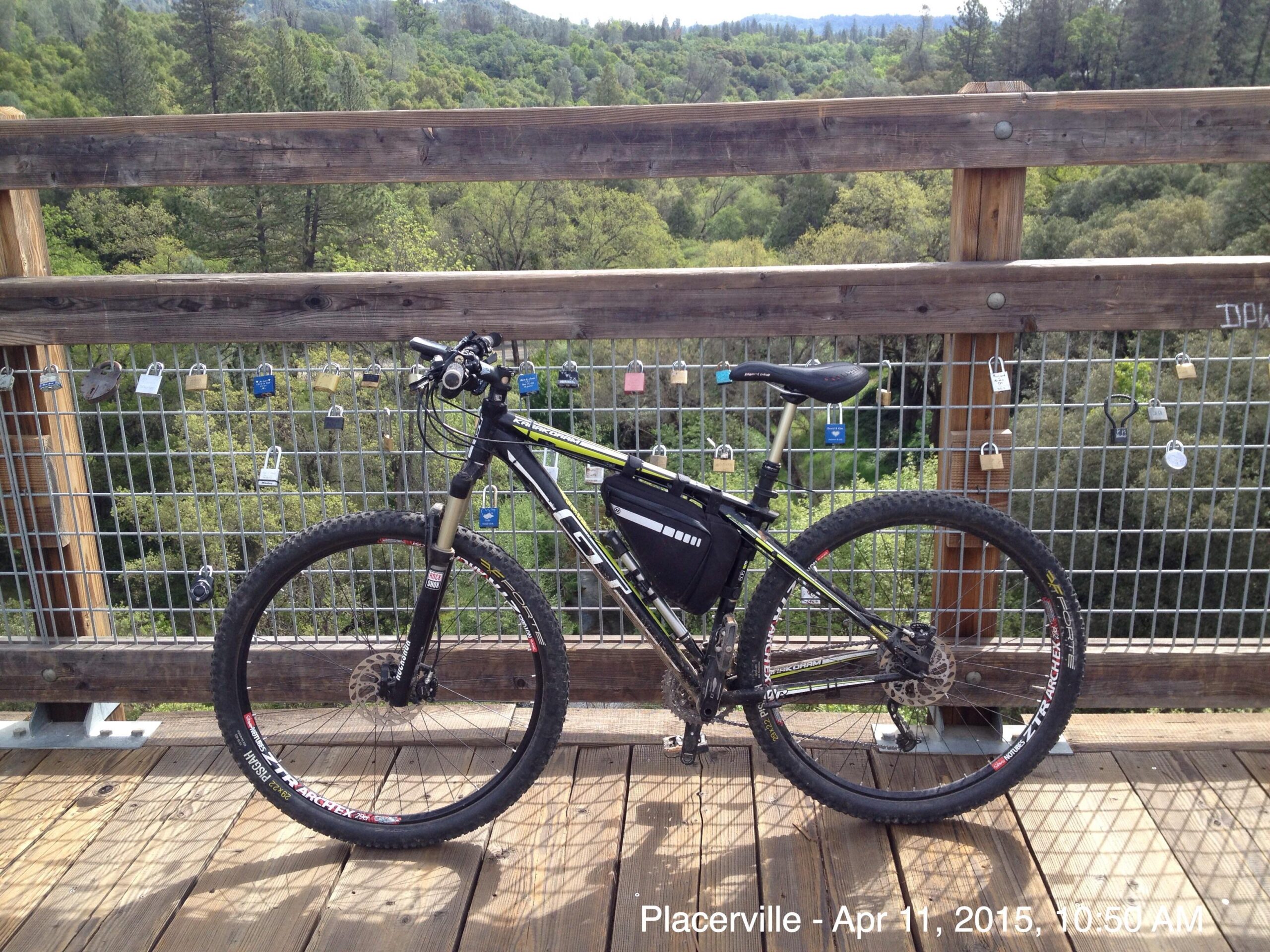A black mountain bike is parked on a wooden deck with a metal railing. The background features lush green trees and hills under a clear sky. Several colorful padlocks are attached to the railing, suggesting a place of significance or memory. El Dorado Trail mountain bike trail.