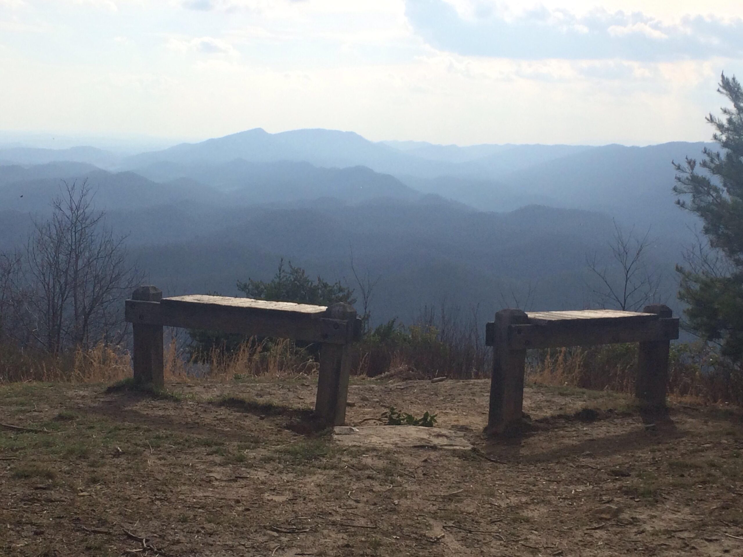 A scenic view from a mountain overlook featuring two wooden benches facing distant rolling hills and a cloudy sky. The foreground shows grassy terrain, while the background displays layers of blue mountains fading into the horizon. Hungry Mother State Park mountain bike trail.