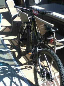 A black mountain bike is parked on a patio, with sunlight casting shadows on the ground. In the background, there are outdoor chairs and a table.