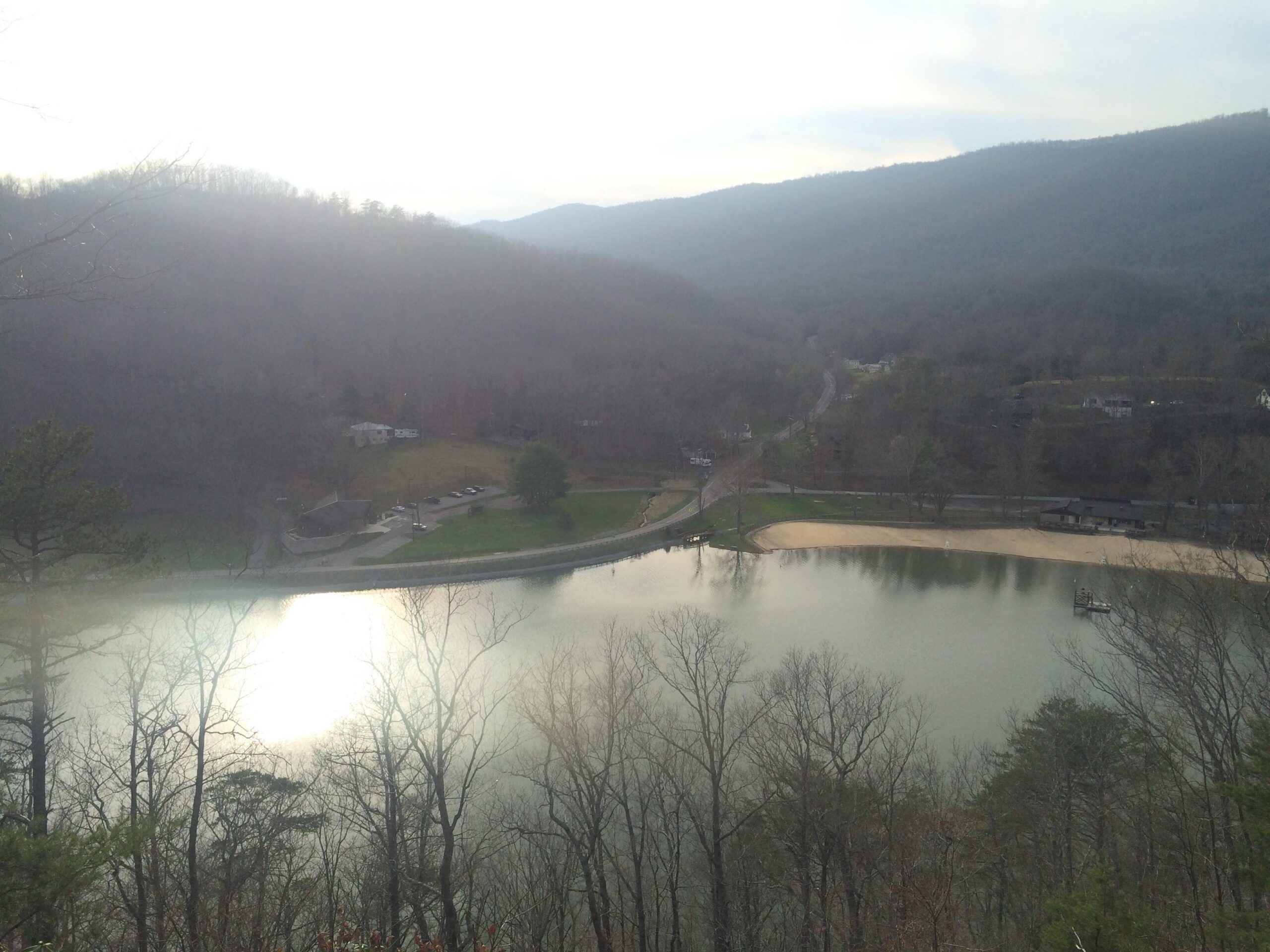 A scenic view of a calm lake surrounded by wooded hills, with soft sunlight reflecting on the water's surface. In the foreground, bare trees are visible, while buildings and a sandy beach are situated along the lake's edge. A winding road can be seen nearby, leading through the landscape. Hungry Mother State Park mountain bike trail.
