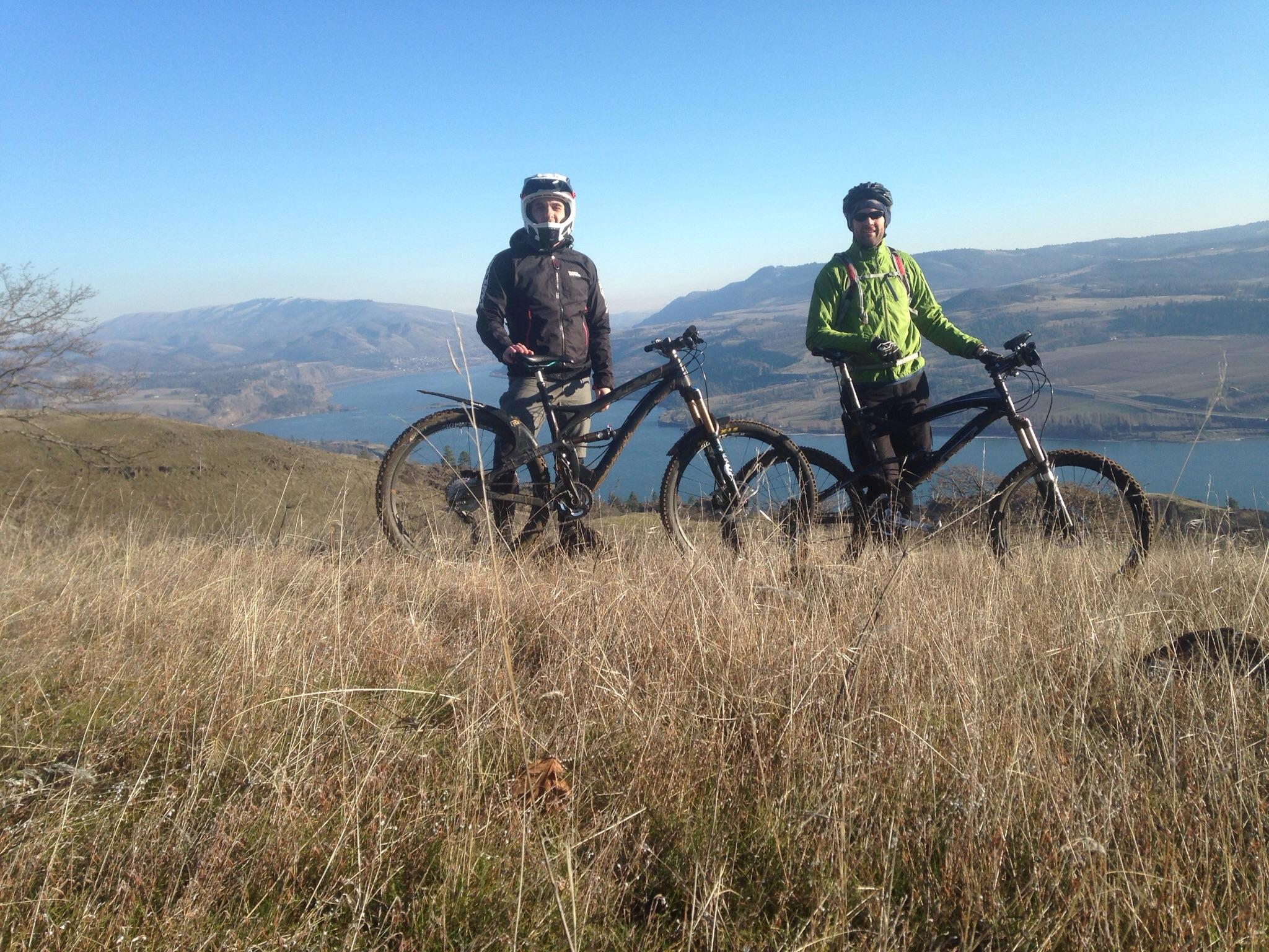 Two mountain bikers stand on a grassy hilltop with bikes beside them, overlooking a scenic landscape featuring a lake and distant mountains under a clear blue sky. One rider is wearing a black jacket and helmet, while the other is dressed in a bright green jacket. Syncline mountain bike trail.