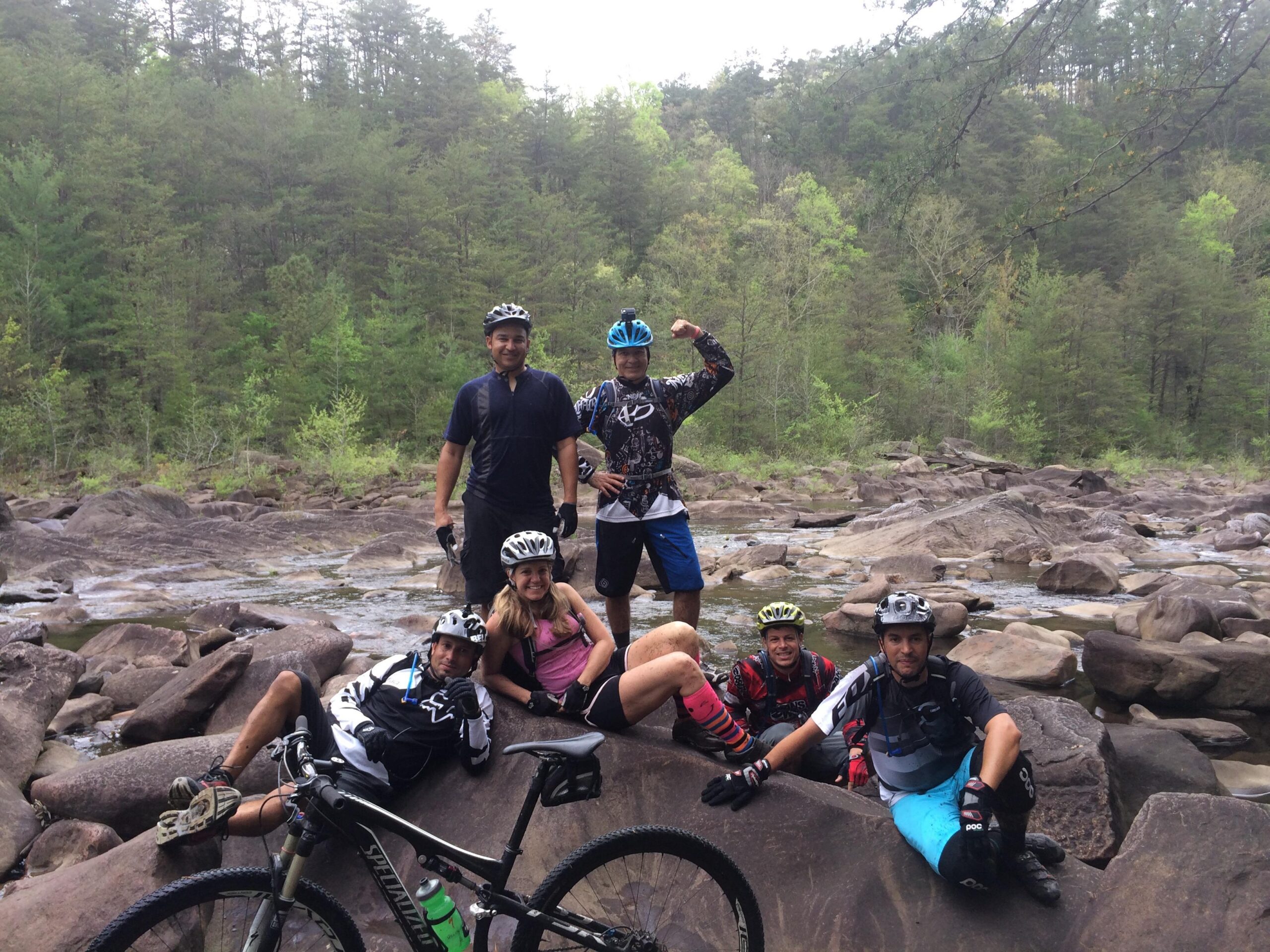 A group of six mountain bikers posing on a rocky riverbank surrounded by lush greenery. Some are casually sitting on the rocks, while others stand, showcasing their bikes and mountain biking gear. The scene captures a spirit of adventure and camaraderie in an outdoor setting. Tanasi Trail System mountain bike trail.