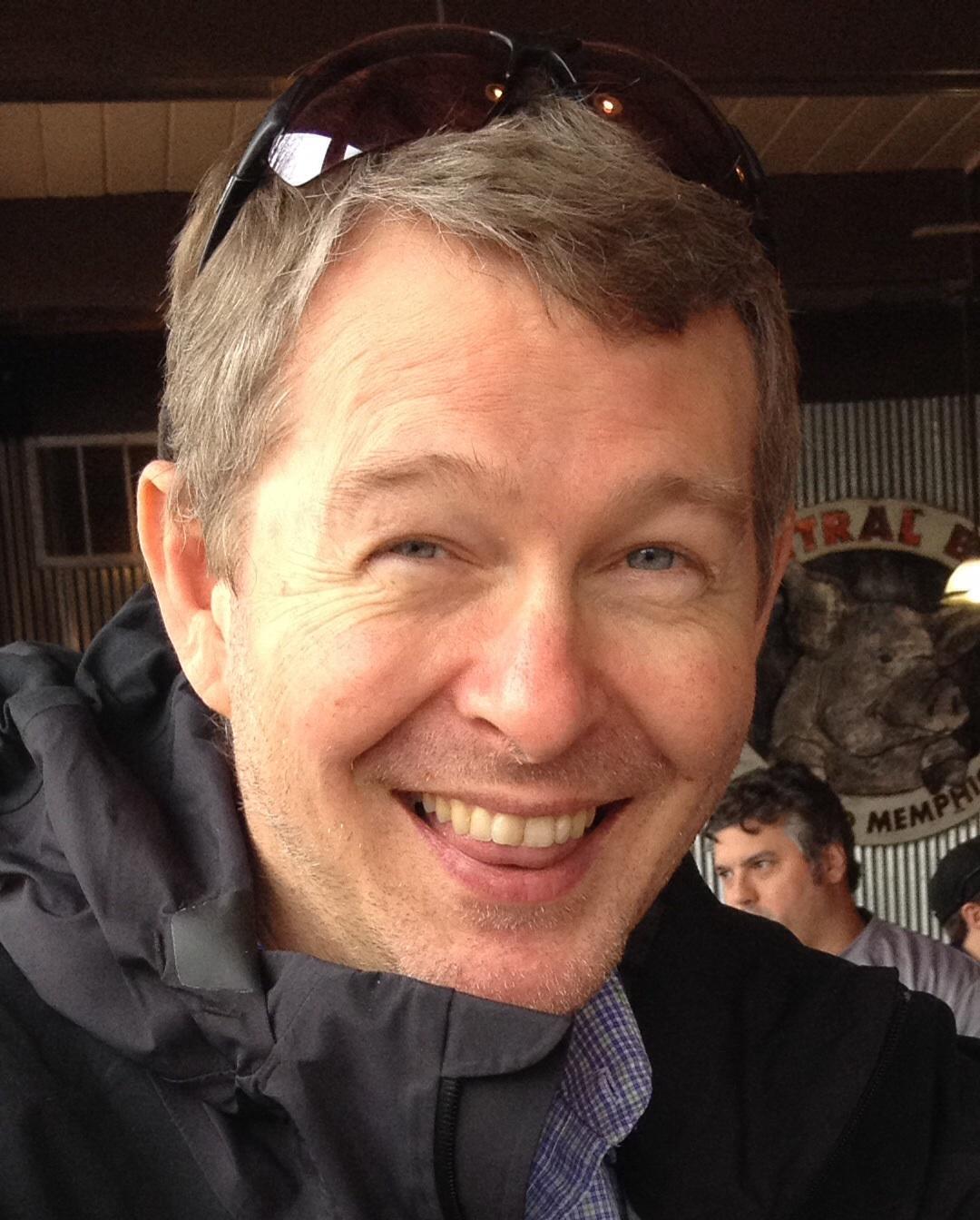 Motobecane NightTrain Bullet: A smiling man with short gray hair, wearing a black jacket, is seen in a casual dining setting. He has sunglasses resting on his head and is sitting at a table with other people in the background. The atmosphere appears relaxed and inviting.