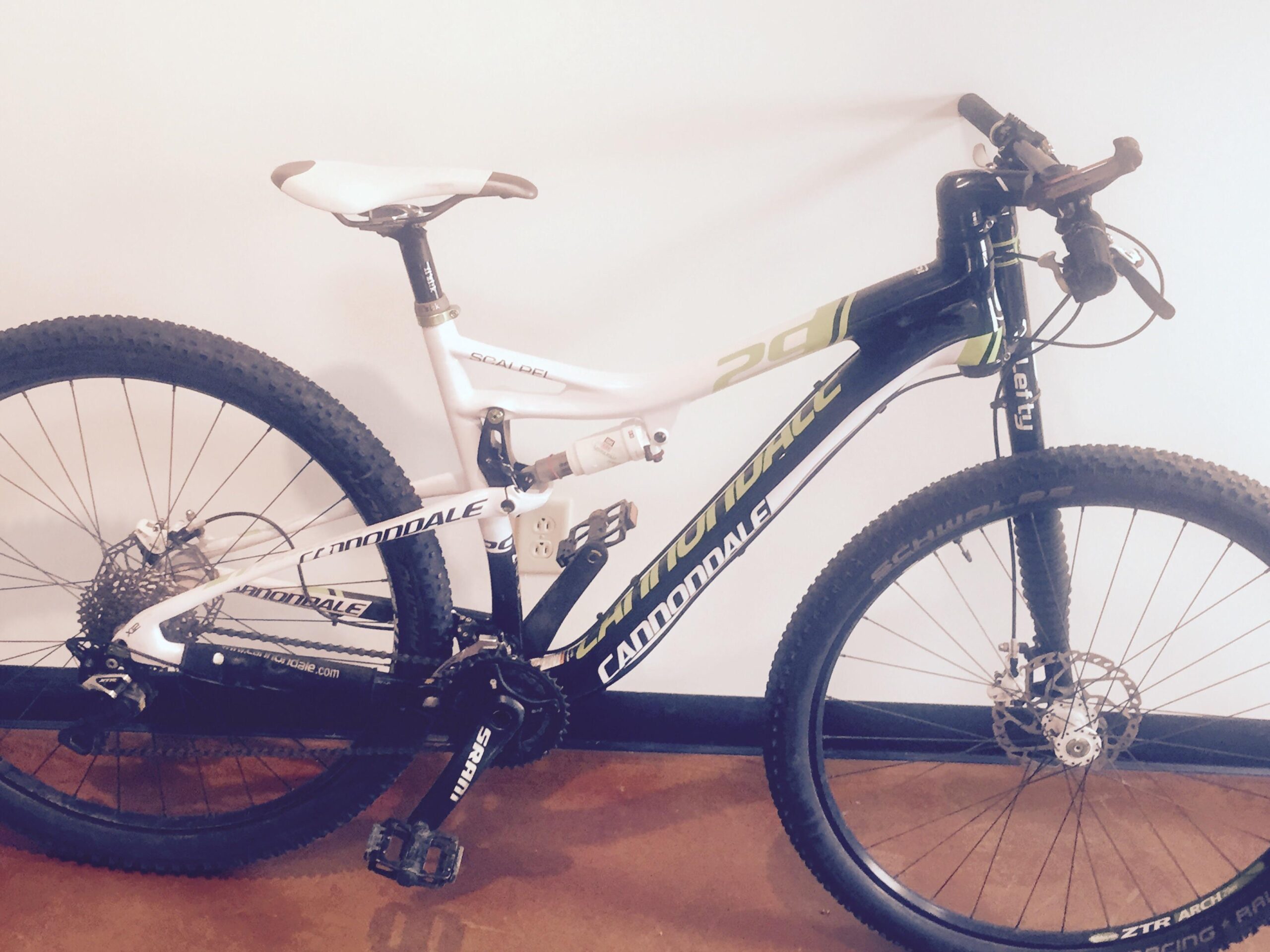 Cannondale Scalpel 29 Carbon 2: Alt text: A Cannondale mountain bike with a white and black frame, featuring large tires and a dual-suspension design, positioned against a plain wall. The bike includes a distinctive lefty fork and is equipped with various gears and brakes.