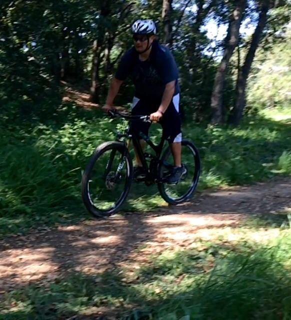 Yeti SB95: A person riding a mountain bike on a dirt path surrounded by greenery and trees on a sunny day. The cyclist is wearing a helmet and casual clothing, focusing on navigating the trail.