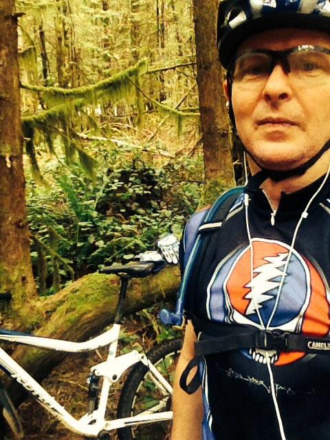 Motobecane NightTrain Bullet: A person wearing a cycling jersey and helmet stands in a forest, with a mountain bike leaned against a fallen log in the background. The scene is lush with greenery, showcasing tall trees and ferns. The cyclist appears to be taking a moment to pause while enjoying the outdoors.