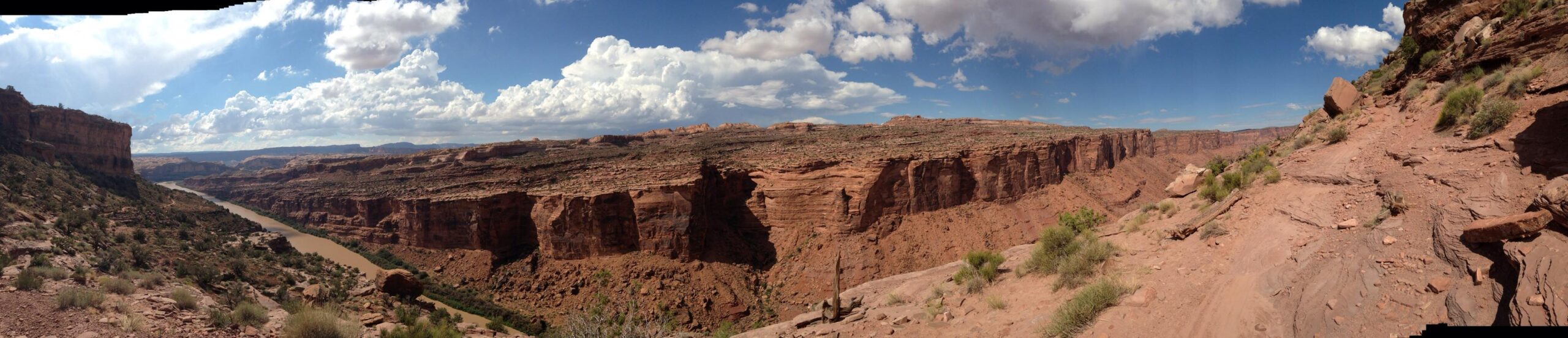 A panoramic view of a rugged canyon landscape featuring steep red rock cliffs, a winding river at the base, and a vast blue sky with scattered white clouds. The terrain is primarily rocky with patches of vegetation, illustrating the natural beauty of the desert environment. Porcupine Rim mountain bike trail.