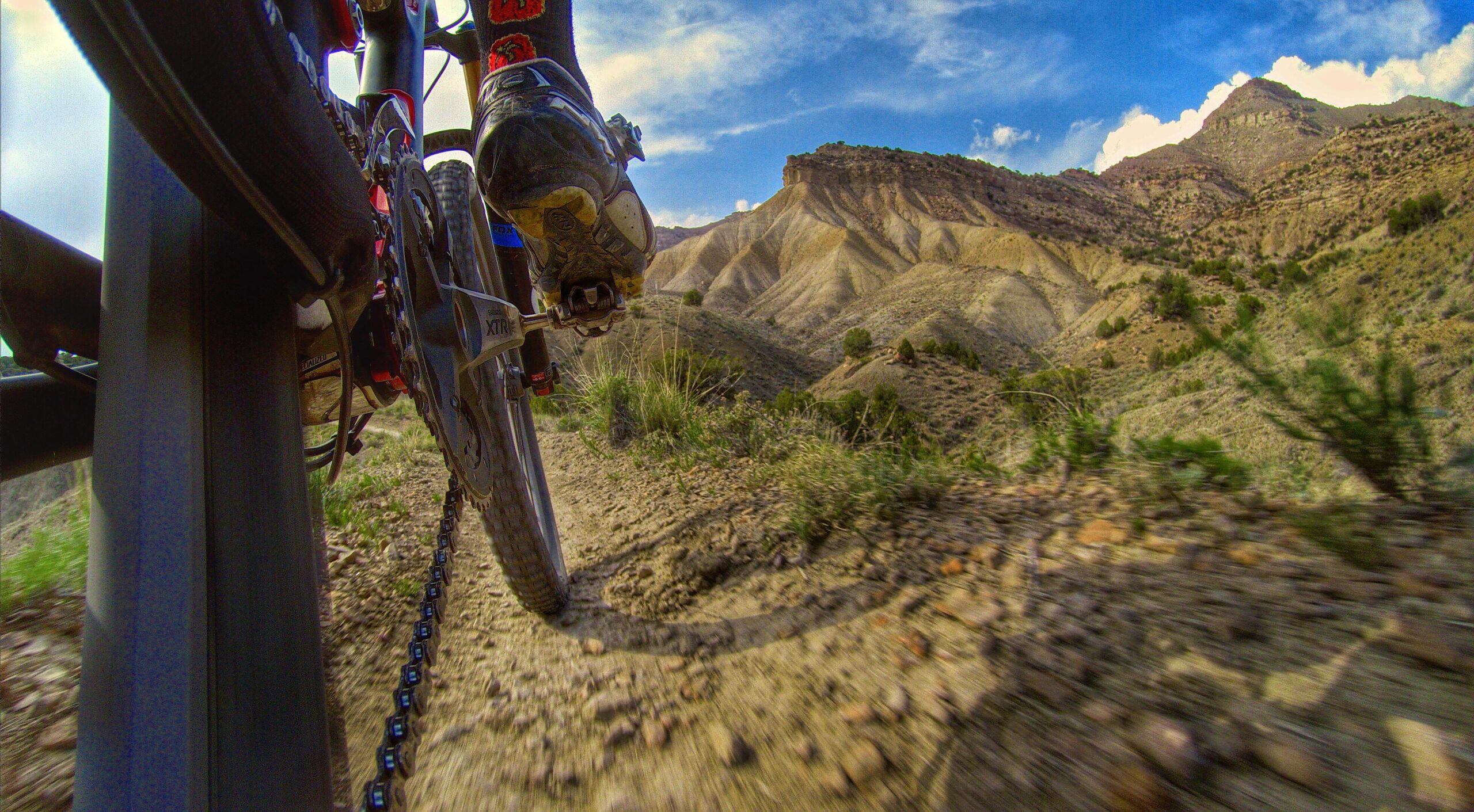 A close-up view of a mountain bike riding along a rugged dirt trail, with the left pedal and a foot in cycling shoes visible. The background features a scenic landscape of rolling hills and mountains under a partly cloudy sky. 18 Road Trails / North Fruita Desert mountain bike trail.