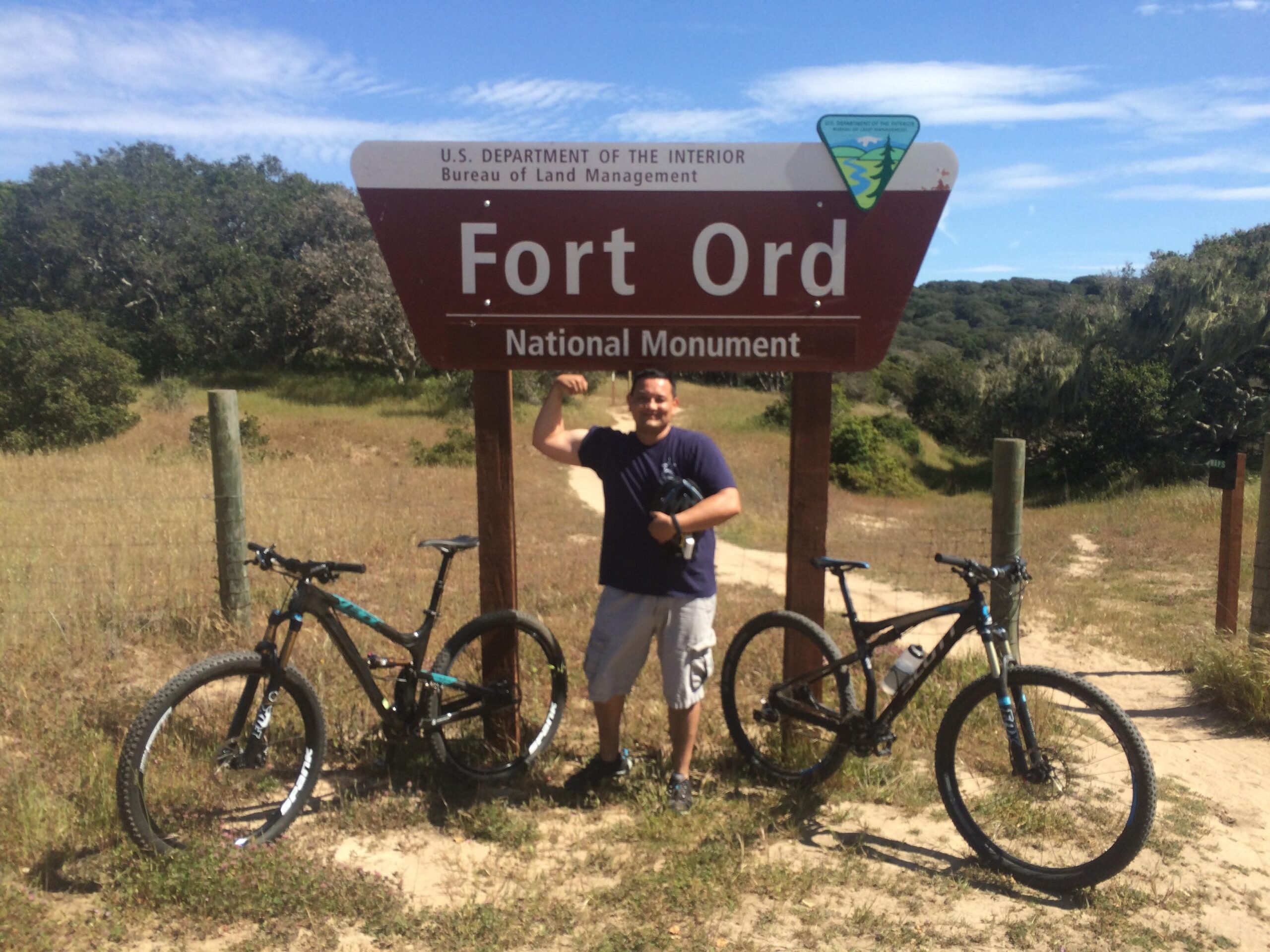 Yeti SB95: A person standing and flexing next to a Fort Ord National Monument sign, with two mountain bikes parked nearby on a dirt path surrounded by grass and trees under a blue sky.
