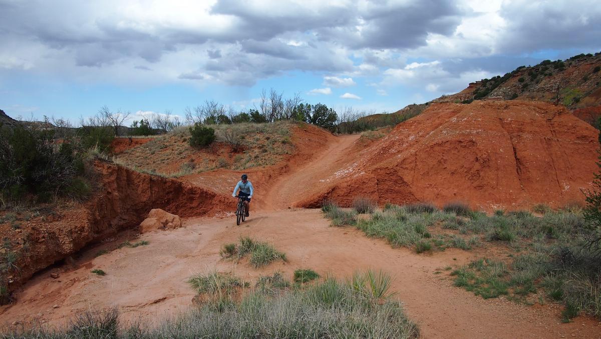 An individual riding a bicycle on a dirt trail surrounded by reddish-brown hills and sparse vegetation under a cloudy sky. Palo Duro Canyon mountain bike trail.