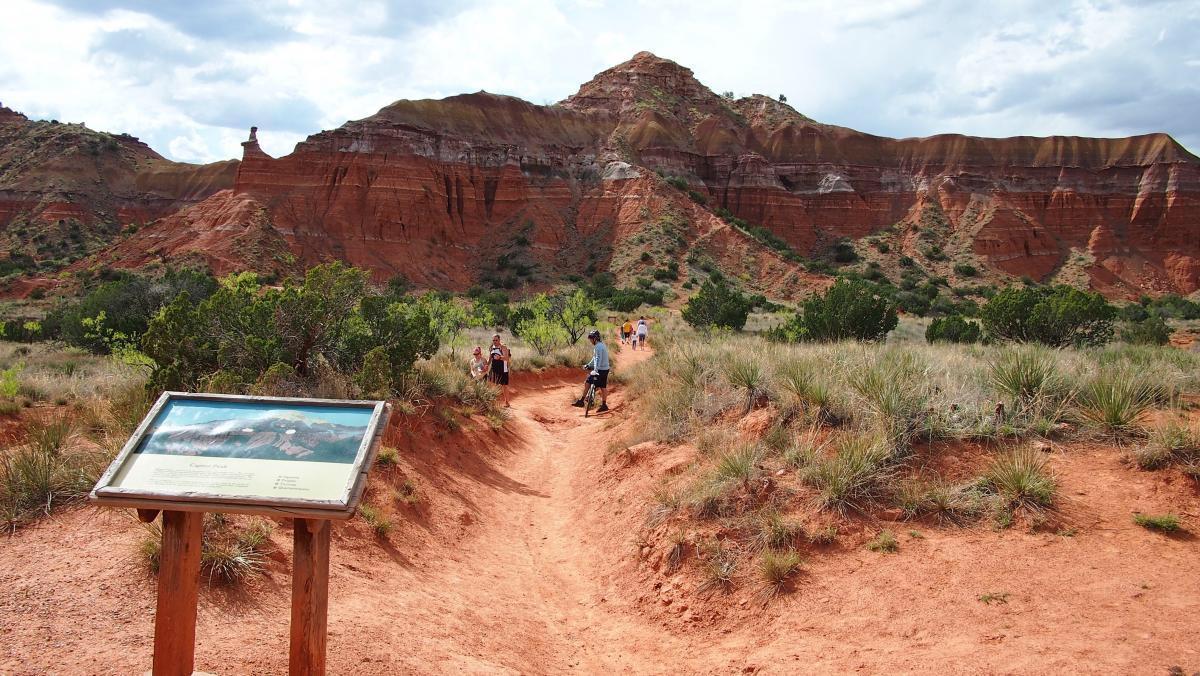 A scenic view of a hiking trail leading through a rugged, red rocky landscape with dramatic cliffs in the background. A trail sign is positioned in the foreground, while a group of trail-goers, including a person on a bicycle, can be seen making their way along the path. The area is filled with sparse vegetation and a partly cloudy sky overhead. Palo Duro Canyon mountain bike trail.
