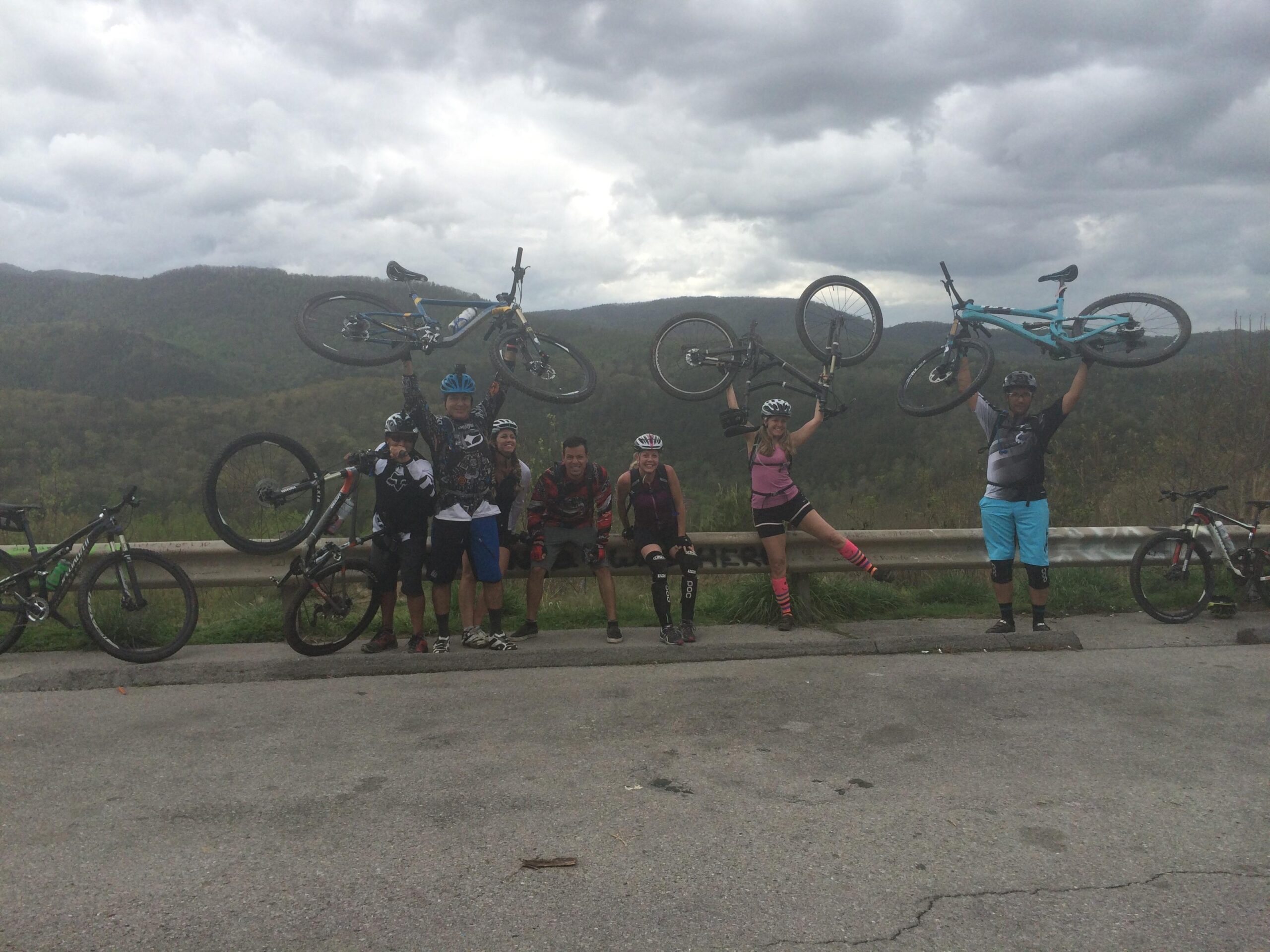 Group of eight mountain bikers celebrating at a scenic overlook, joyfully raising their bikes overhead. The backdrop features rolling green hills under a cloudy sky, with several bicycles resting against a guardrail in the foreground. Tanasi Trail System mountain bike trail.