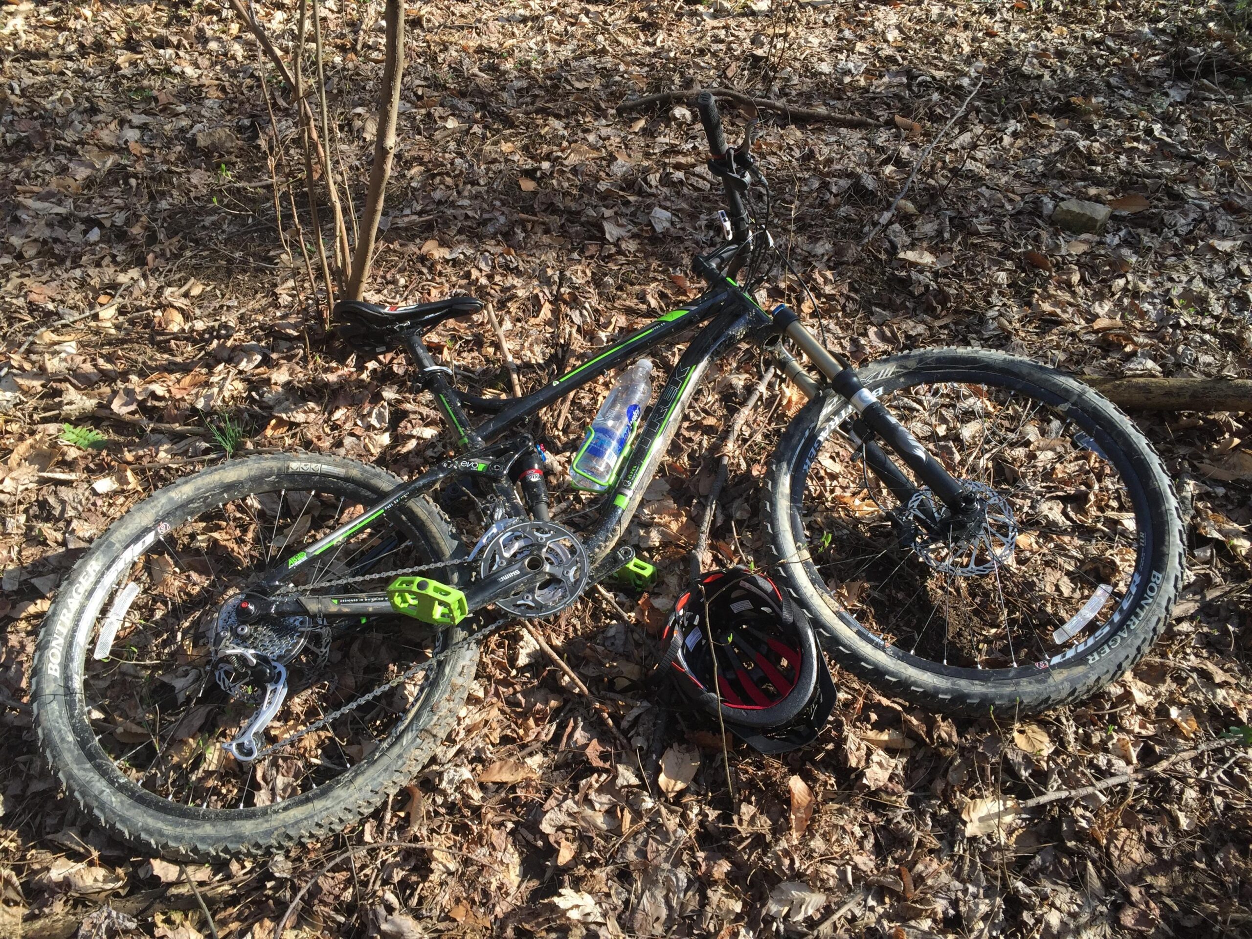 Trek Fuel EX 9: A mountain bike lying on the ground in a forested area, surrounded by dry leaves. A helmet is positioned near the bike, which has a black and green frame and various components visible, including the wheels and chain. Sunlight filters through the trees, casting shadows on the forest floor.