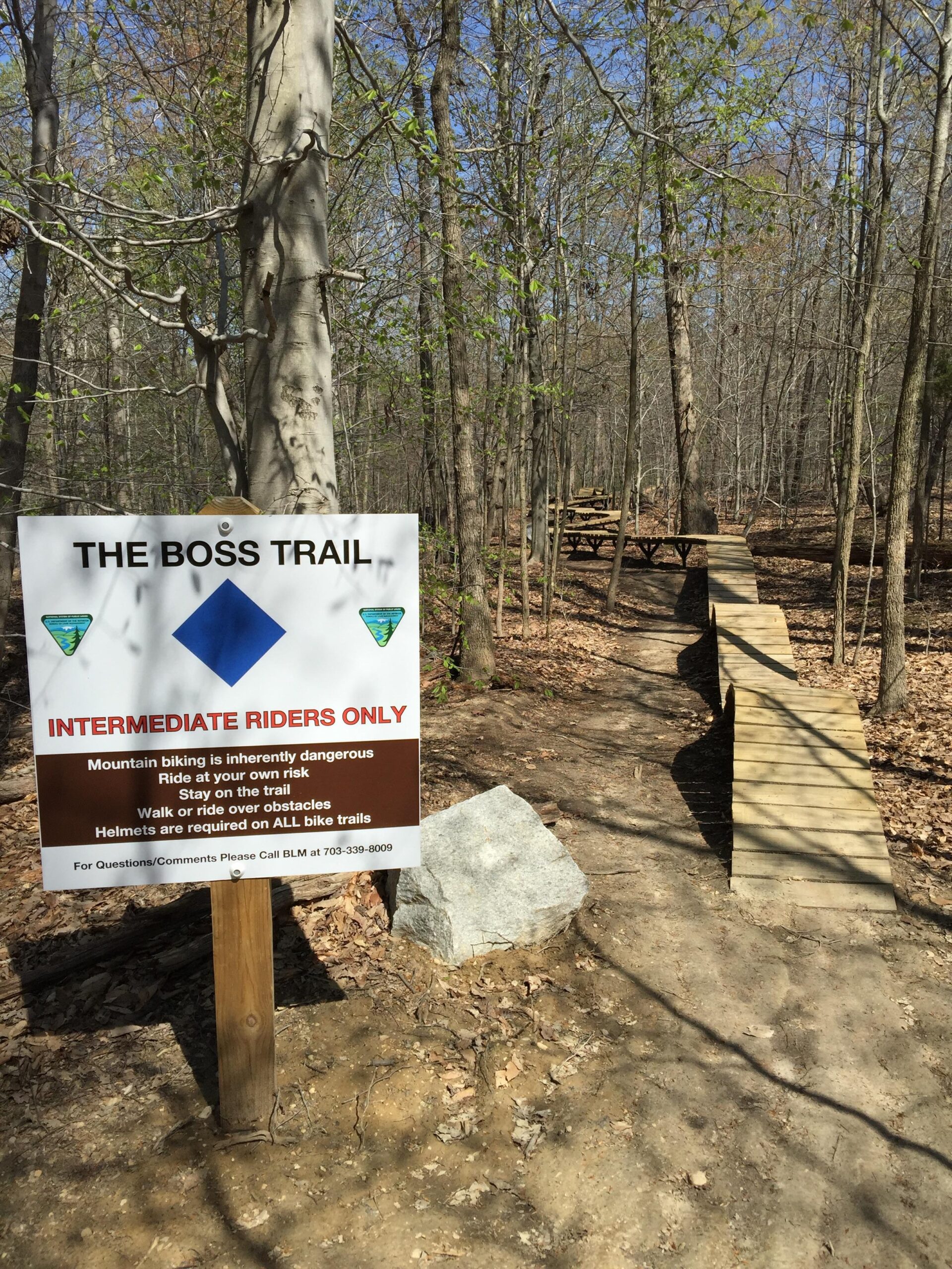 Sign indicating "The Boss Trail" for intermediate mountain bikers, highlighting safety warnings and guidelines. The trail is surrounded by trees and features a wooden pathway leading into the woods. Meadowood mountain bike trail.
