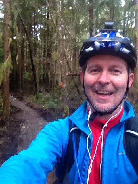 Motobecane NightTrain Bullet: A man wearing a cycling helmet and blue jacket smiles for a selfie while biking on a wooded trail. The background features trees and a dirt path, indicating a forested area for mountain biking.