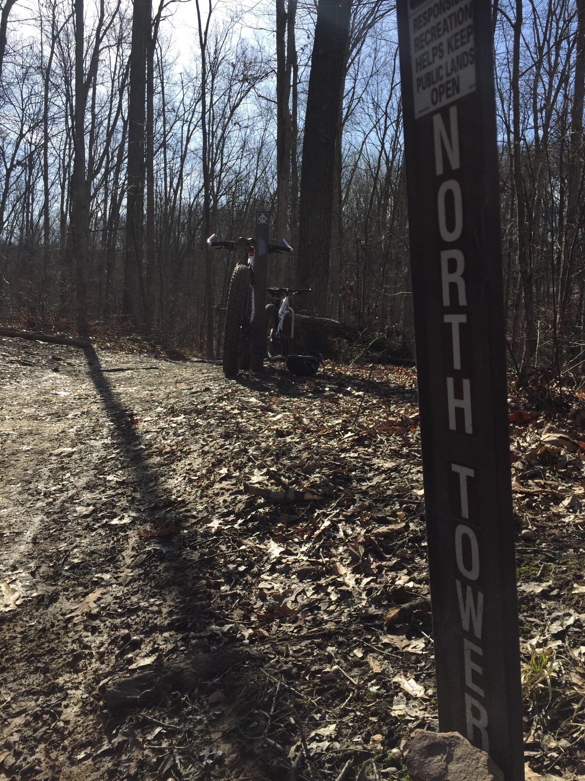 A trail sign reading "NORTH TOWER" stands in a wooded area, with a dirt path covered in fallen leaves and twigs. A bicycle with wide tires is parked nearby, and the sunlight filters through the trees, casting long shadows on the ground. Brown County Park mountain bike trail.