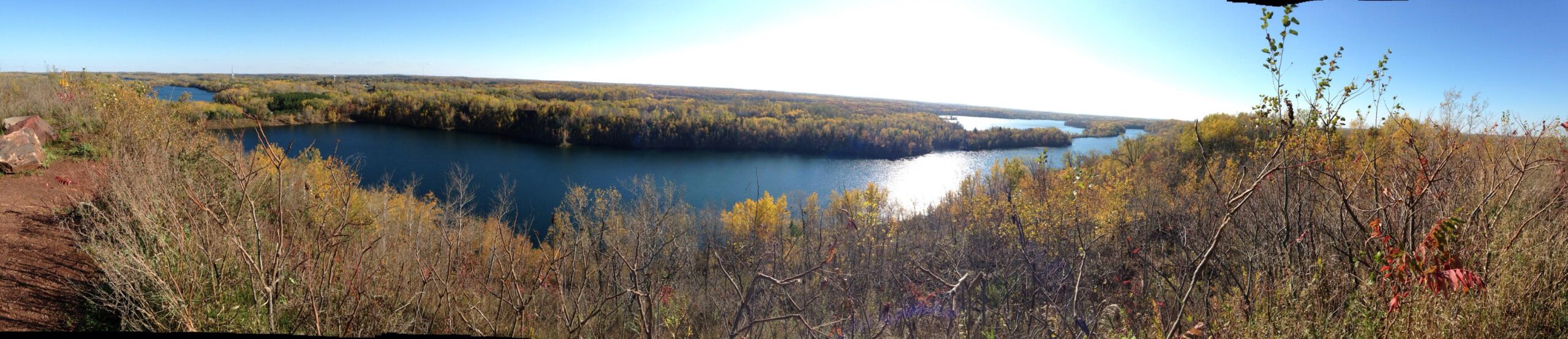 A panoramic view of a serene landscape featuring a winding river surrounded by lush autumn foliage. The scene captures vibrant yellow and orange leaves on trees, contrasting against a clear blue sky. The foreground includes natural vegetation and rocks, while the background showcases rolling hills and additional bodies of water. Cuyuna Lakes mountain bike trail.