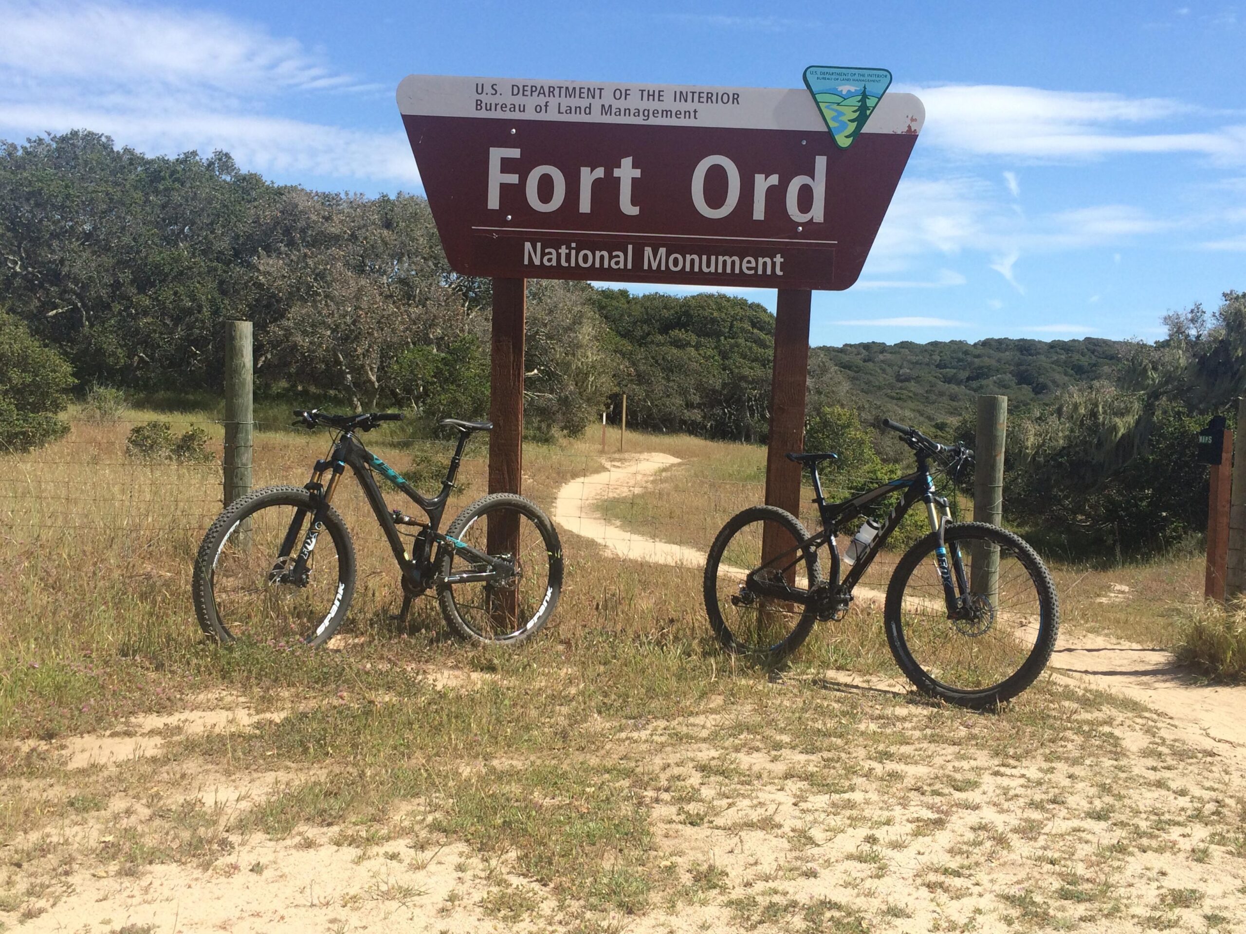 Yeti SB95: Bicycles parked near a sign for Fort Ord National Monument, surrounded by grass and trees, with a dirt path visible leading into the park. The sky is clear with some clouds.