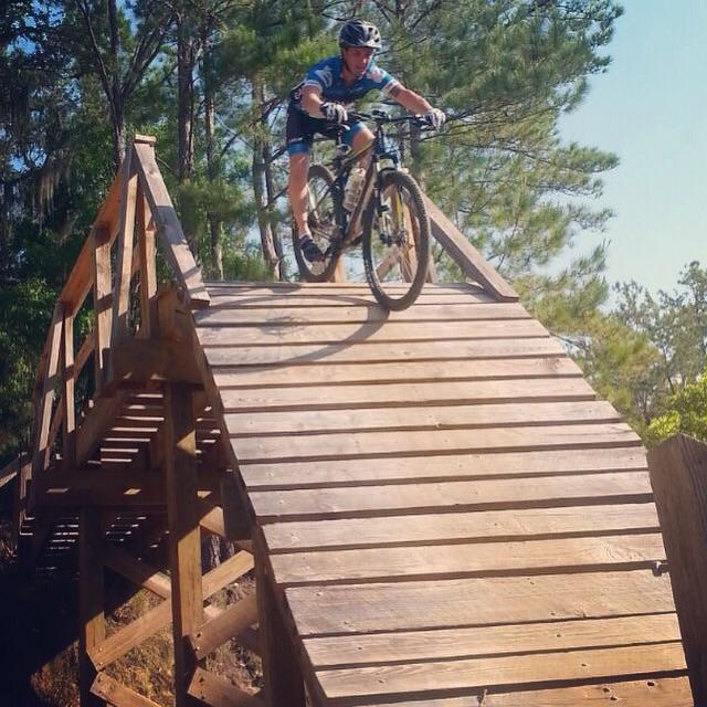 A mountain biker in a blue jersey and black shorts performs a jump off a wooden ramp surrounded by trees, showcasing skill and balance. Santos mountain bike trail.