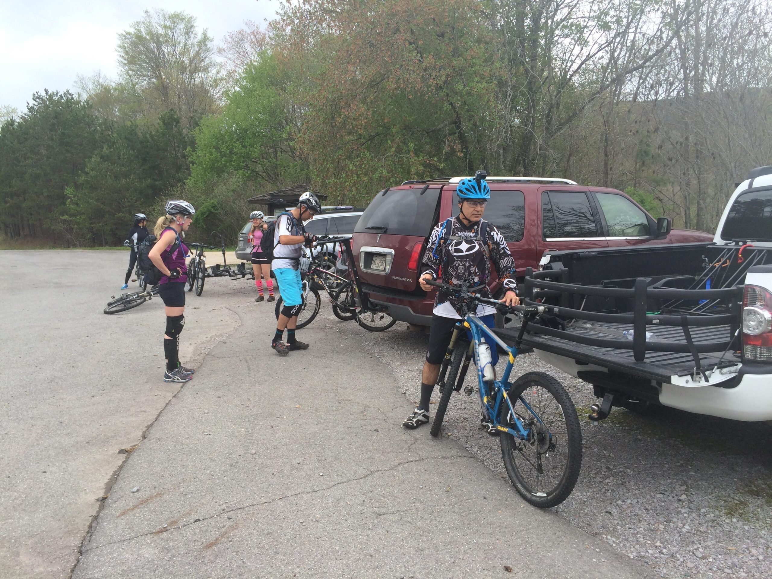 A group of mountain bikers preparing for a ride in a parking area. Some are adjusting their gear while others are getting their bikes ready. The scene includes several parked cars, greenery in the background, and a mix of riders wearing helmets and protective gear. The atmosphere appears casual and focused on cycling. Tanasi Trail System mountain bike trail.