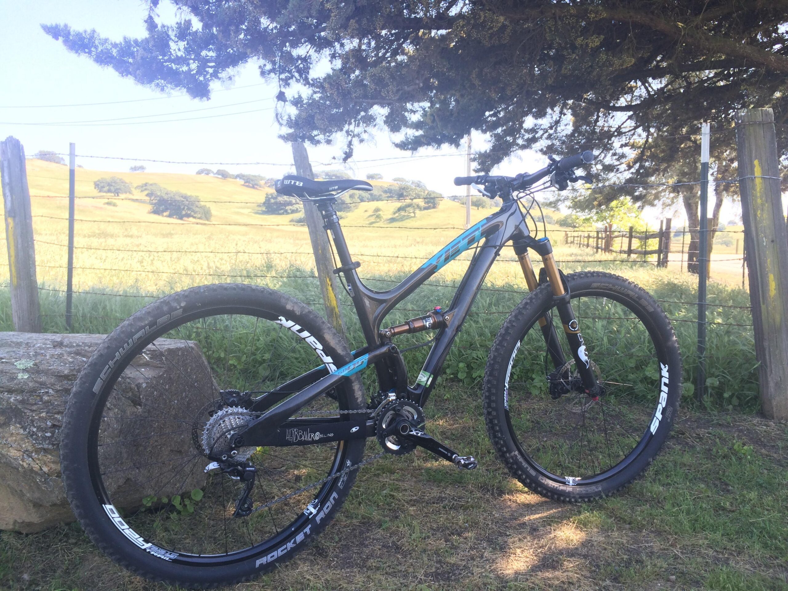 Yeti SB95: A high-performance mountain bike parked on grass near a stone, with a scenic rural landscape featuring rolling hills and trees in the background.