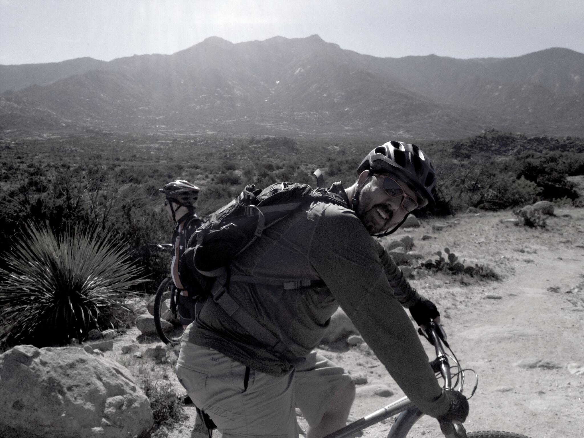 A man wearing a helmet and sunglasses smiles while riding a mountain bike along a dirt trail. He is dressed in outdoor gear and has a backpack on. In the background, a rugged mountainous landscape and vegetation are visible under a clear sky. The scene conveys a sense of adventure and outdoor recreation. 50-year Trail / Golder Ranch mountain bike trail.