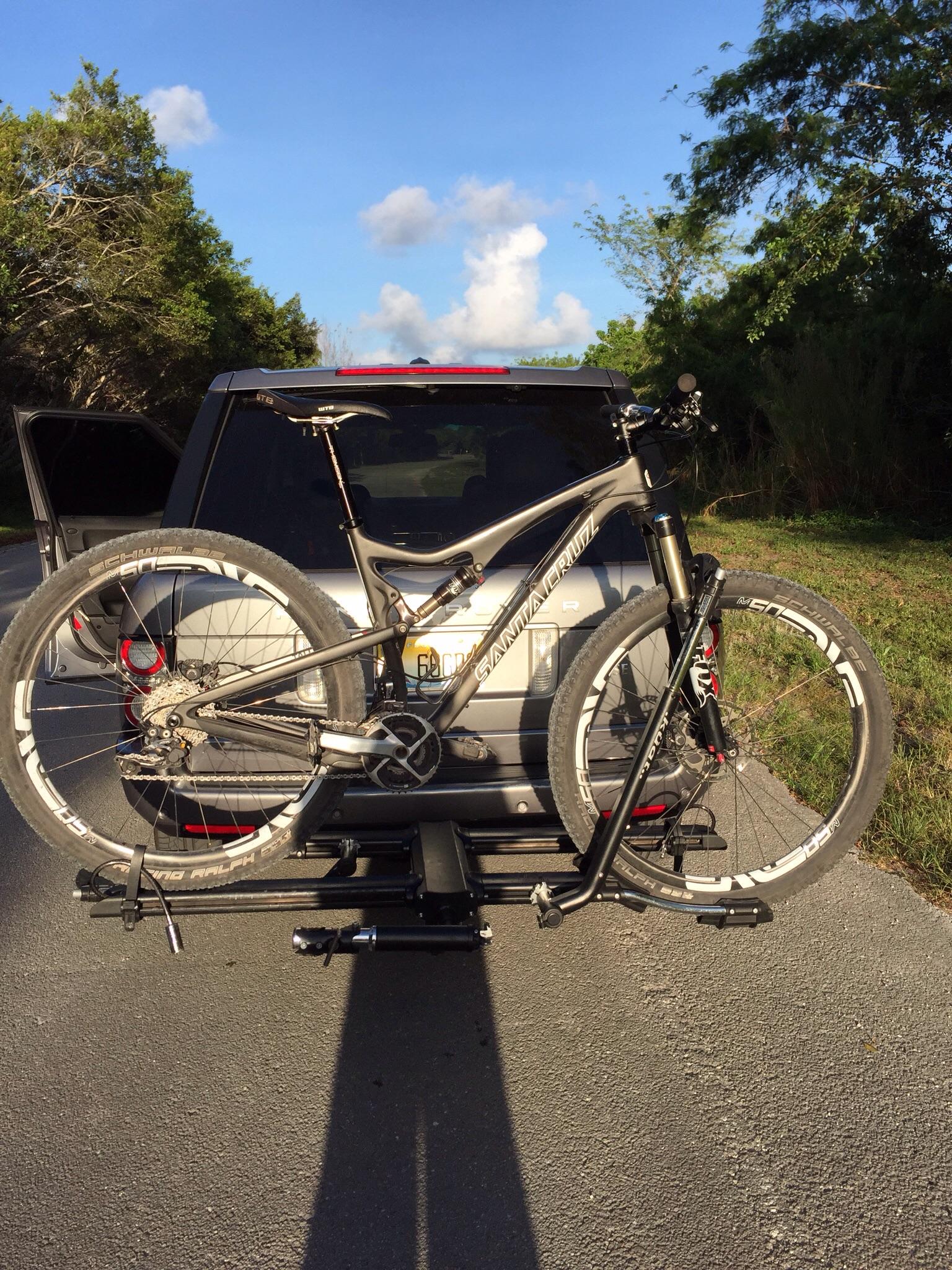 Santa Cruz Tallboy Carbon: A mountain bike secured to a bike rack on the back of a vehicle, parked on a road surrounded by greenery and a blue sky with scattered clouds. The vehicle's trunk is open, indicating preparation for a biking trip.