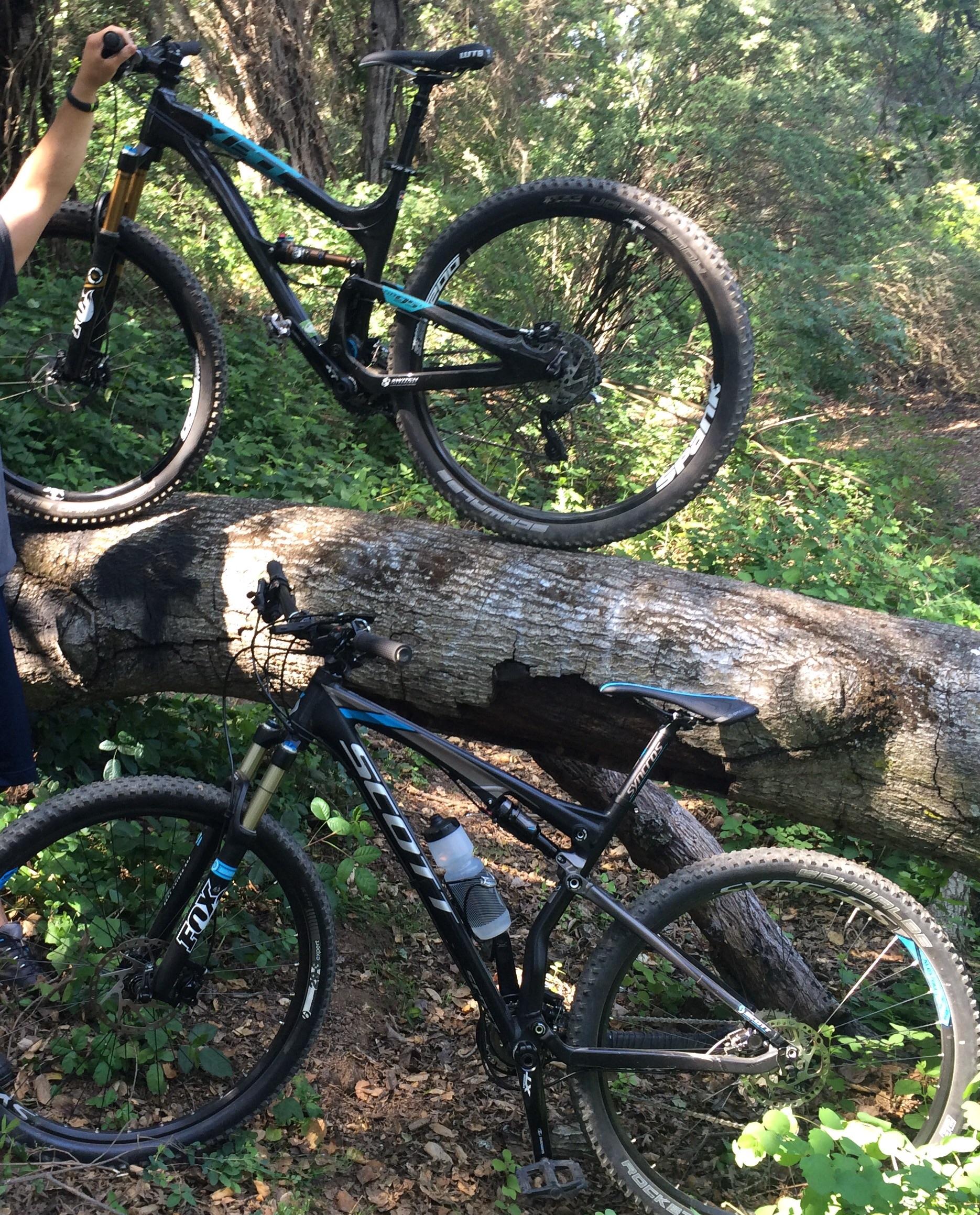 Yeti SB95: Alt text: Two mountain bikes are positioned alongside a fallen tree in a wooded area. One bike is held up by a person, while the other rests on the ground, partially obscured by foliage.