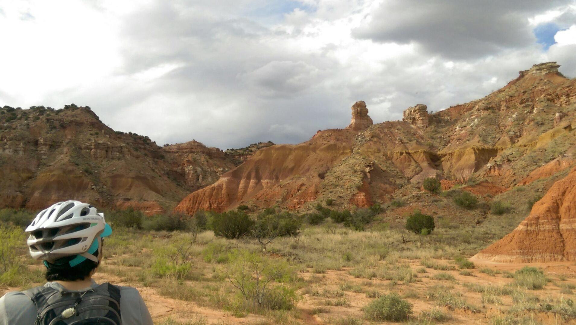A person wearing a white bicycle helmet is seen from behind, looking out over a landscape of colorful, rocky hills and cliffs. The terrain features various shades of red, orange, and brown, with sparse vegetation and a cloudy sky overhead. Palo Duro Canyon mountain bike trail.