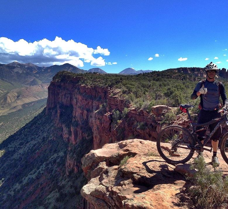 A mountain biker stands on the edge of a rocky cliff, giving a thumbs-up gesture. The background features a breathtaking landscape of rolling hills and mountains under a bright blue sky with fluffy white clouds. Lush greenery covers the terrain below, adding to the scenic view. The Whole Enchilada mountain bike trail.
