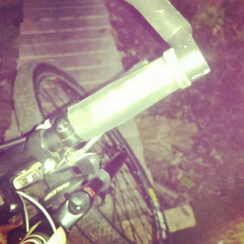 An overhead view of a bicycle handlebar at night, showing the grips, brake lever, and part of the bike frame, with a blurred background of stones and vegetation. Mb Johnson Park mountain bike trail.