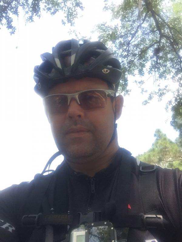 A person wearing a bicycle helmet and sunglasses, standing outdoors in a wooded area. They are dressed in a black jersey and have a camera mounted on their chest. Sunlight filters through the trees in the background. Halpatiokee mountain bike trail.
