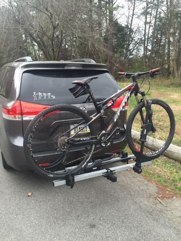A mountain bike secured on a hitch-mounted bike rack attached to the back of a gray minivan, parked in a wooded area. The rear of the van features playful stick figure decals. Powhite Park mountain bike trail.