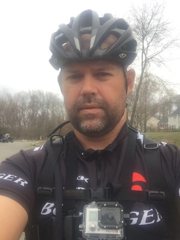A close-up selfie of a man wearing a cycling helmet and a black cycling jersey with a logo. He is equipped with a camera mounted on his chest harness. The background shows a muted, outdoor setting with trees and a residential area. Powhite Park mountain bike trail.