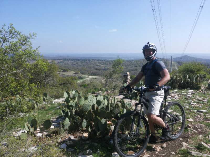 A person wearing a helmet and cycling gear stands beside a mountain bike on a rocky path. The background features a scenic view of rolling hills, trees, and cacti under a clear blue sky. Power lines stretch across the landscape, indicating the outdoor setting. Flat Rock Ranch mountain bike trail.