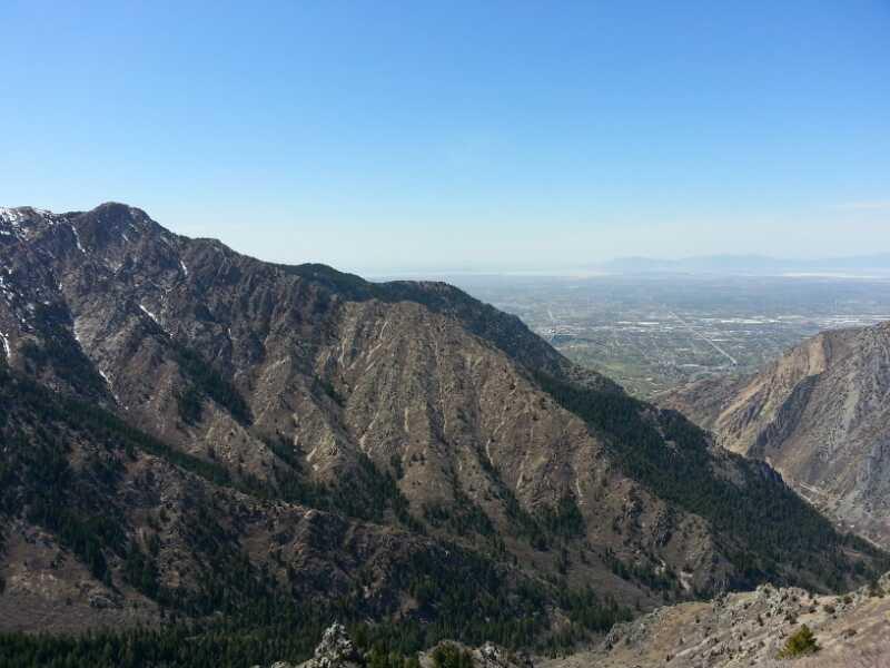 A panoramic view of rugged mountains with a mixture of rocky terrain and patches of greenery, under a clear blue sky. In the distance, a sprawling landscape can be seen, including valleys and small urban areas. The scene captures the natural beauty and serene atmosphere of the mountainous region. Coldwater Canyon Overlook mountain bike trail.