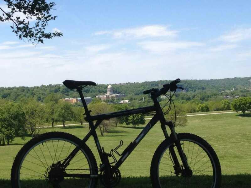A silhouette of a mountain bike is positioned in the foreground, overlooking a green landscape with rolling hills. In the background, a building with a dome is visible among trees, under a blue sky with scattered clouds. Capital View mountain bike trail.