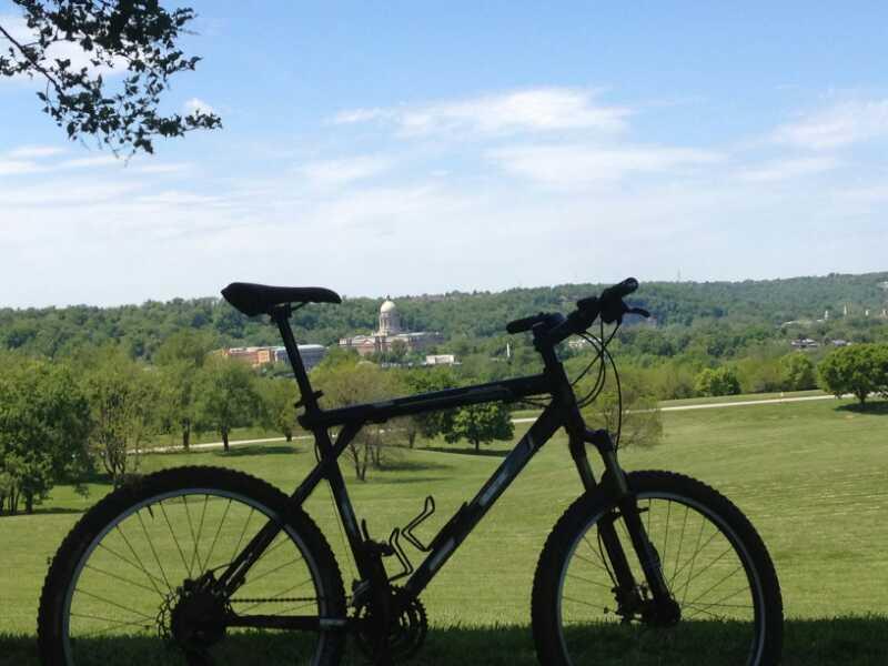 A silhouette of a mountain bike in the foreground, with a lush green landscape and rolling hills in the background. A clear blue sky with a few clouds is overhead, and a building with a dome is visible in the distance among the trees. Capital View mountain bike trail.