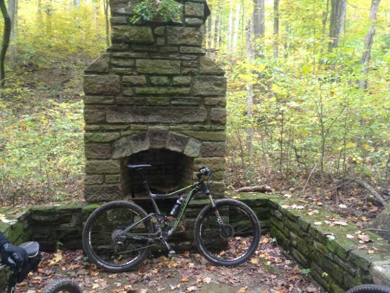 A mountain bike parked next to a stone fireplace in a wooded area with autumn foliage. The scene reveals a serene forest background with trees displaying yellow and green leaves. Brown County Park mountain bike trail.
