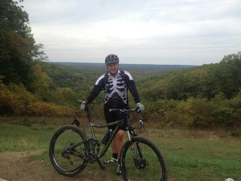 A person wearing a skeleton-themed cycling jersey stands beside a mountain bike on a grassy hillside. In the background, a panoramic view of a forested landscape stretches out under a cloudy sky. Brown County Park mountain bike trail.
