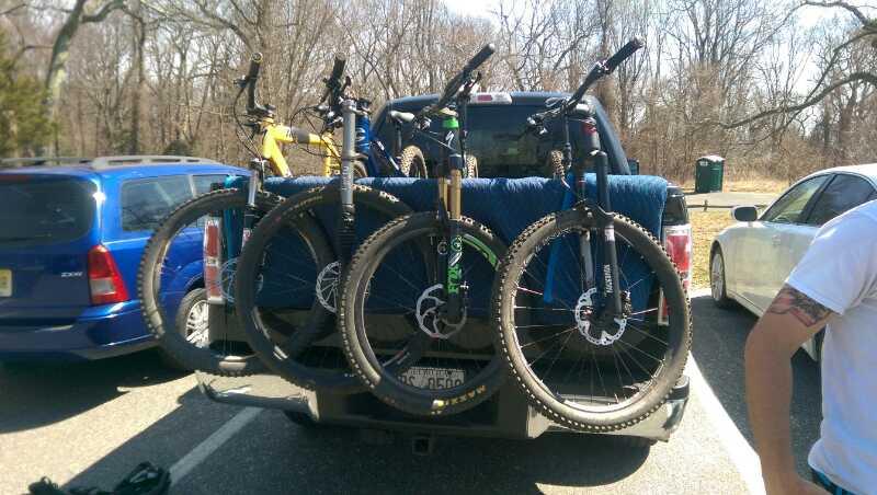 Three mountain bikes mounted on the back of a pickup truck, with a blue blanket draped over the tailgate. A blue vehicle is parked to the left, while a white car is visible on the right. The background features bare trees, indicating early spring or late winter. Hartshorne Woods Park mountain bike trail.