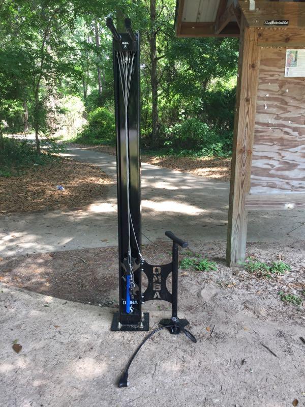 A black outdoor fitness station with cables for resistance training, featuring a dual pulley system, located in a wooded area near a pathway. It includes a footplate for stability. Santos mountain bike trail.