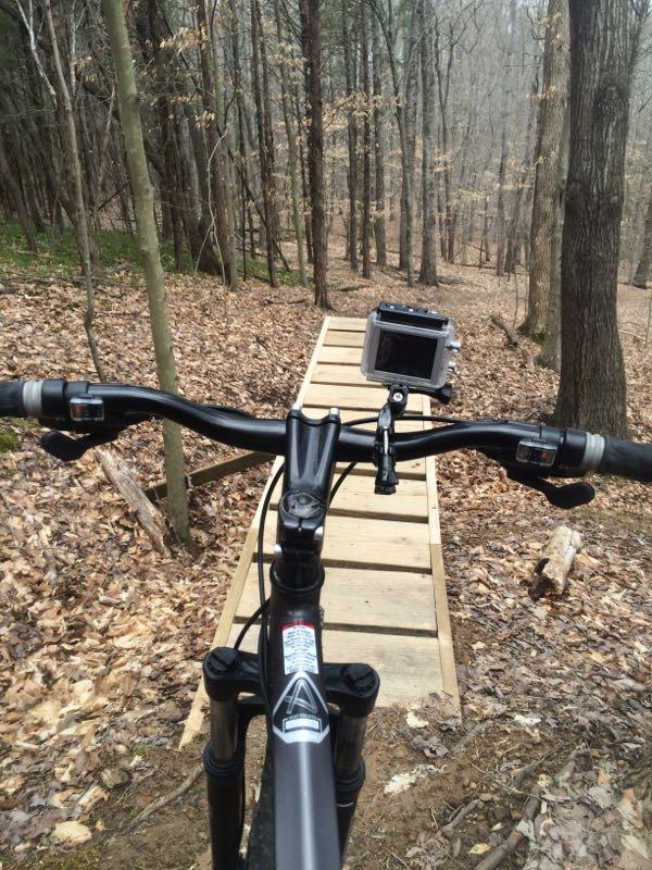 View from the handlebars of a mountain bike, showing a wooden ramp ahead in a wooded area with sparse leaves on the ground. A camera is mounted on the handlebars, pointing down the ramp. Green River mountain bike trail.