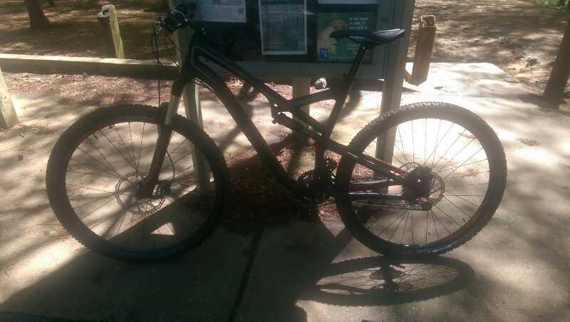 A black mountain bike parked on a paved surface near a bulletin board in a wooded area, with sunlight filtering through the trees. Sope Creek mountain bike trail.
