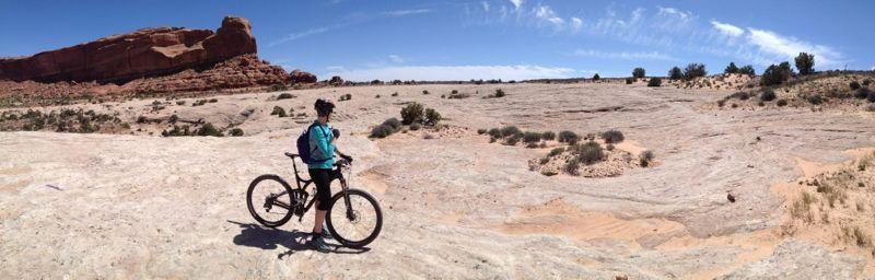 A person riding a mountain bike stands on a rocky, arid landscape with sparse vegetation, under a clear blue sky with wispy clouds. In the background, a large red rock formation is visible, highlighting the area's rugged terrain. Navajo Rocks mountain bike trail.